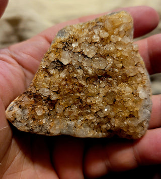 A close up of a hand holding a Merchant's Stone, displaying a layer of golden citrine crystals that glisten under the light, creating a sparkling, faceted appearance.