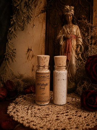 Two small glass bottles with cork stoppers containing loose powder or granules, placed on a crocheted doily in a moody, vintage-style setting with dried flowers, roses, and a figurine in the background. The labels read 'ALVARADO Apothecary and 'All Night Long'.