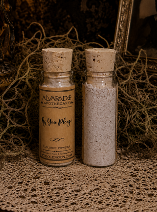 Two small glass apothecary bottles with cork stoppers, one labeled 'Alvarado Apothecary As You Please' and the other plain, filled with light gray powder, displayed on a lace surface with dried moss in the background.