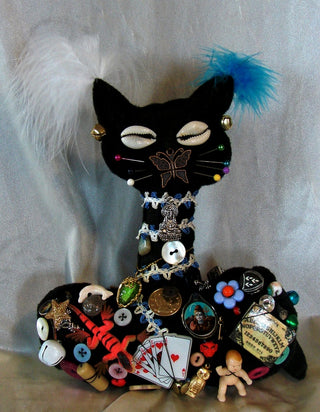 A handmade black felt cat pincushion decorated with buttons, pins, feathers, and small embellishments, set against a light gray fabric background.