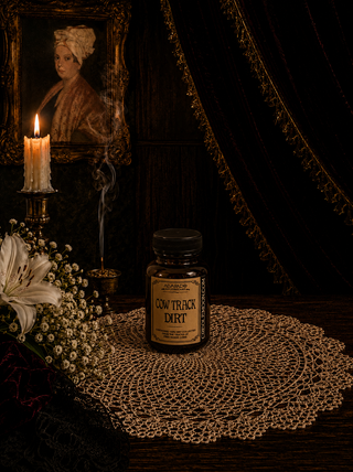 Jar of 'Cow Track Dirt' on a doily with a candle and portrait in the background