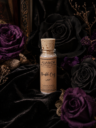 A small glass bottle with a cork stopper and a tan label reading 'ALVARADO Apothecary Double Cross' stands on dark velvet fabric, surrounded by black and deep purple roses and dried botanicals.