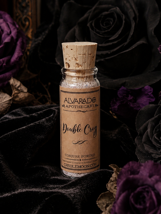 A small glass bottle with a cork stopper and a tan paper label reading 'Alvarado Apothecary Double Cross Powder' is centered against a dark velvet background with black and purple roses.