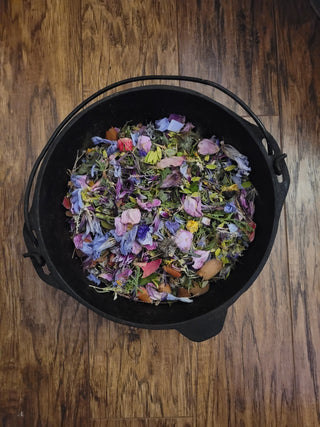 A black cast iron cauldron filled with a colorful mixture of dried flowers, petals, and botanicals, photographed from above on a rustic wooden surface.