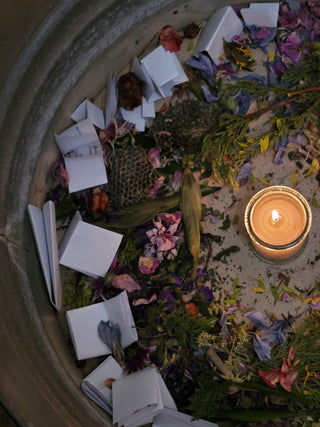 A circular stone container filled with scattered white folded paper pieces, dried flowers, green leaves, and a lit candle in the center.