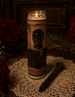 A lit glass prayer candle with a sepia-toned label featuring a black portrait illustration and the text 'JOHN HENRY' near the bottom, styled on a lace doily with a rusted nail and dark red roses around it.