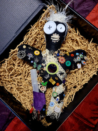 Decorative ju ju doll with intricate embellishments in a box filled with crinkle paper.
