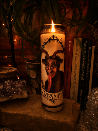 A lit white pillar candle with a dark goat-headed demon illustration and the word 'KRAMPUS' on the front, standing on a stone surface with books, crystals, and plants in a dimly lit setting.