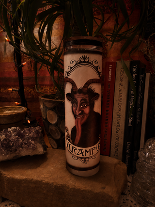 A tall white prayer candle featuring a dark illustration of a horned demon-like face with the word 'KRAMPUS' at the bottom, displayed on a stone surface with books, crystals, and plants in a dimly lit setting.