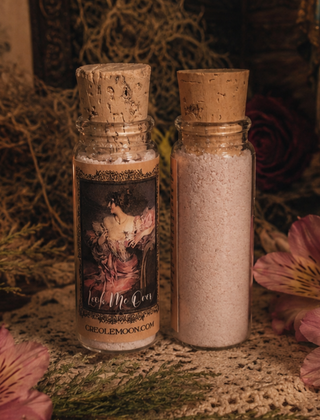Two glass bottles with cork stoppers containing bath salts, displayed on a textured surface with dried flowers and greenery. One bottle has a decorative label reading 'Look Me Over' and 'CREOLEMOON.