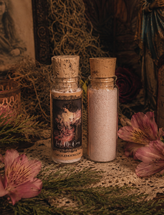 Two small glass bottles with cork stoppers sit on a lace surface surrounded by dried flowers and greenery. The left bottle has a dark label reading "Look Me Over;" the right bottle contains a pale pink powder.
