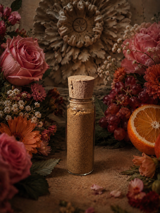 A small clear glass bottle with a cork stopper filled with brown powdered palo santo, styled among pink and orange flowers, grapes, dried botanicals, and an orange slice on a rustic surface.