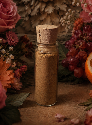 A clear glass bottle with a cork stopper filled with fine brown palo santo powder, styled among dried flowers, red grapes, and an orange slice on a rustic brown surface.