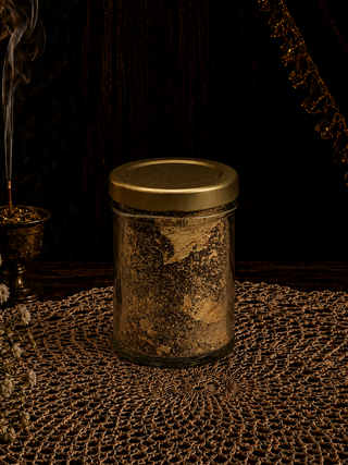 Glass jar of prosperity dust with a gold lid on a textured surface with a dark background