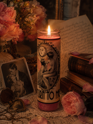 Candle with vintage design and text, surrounded by flowers and books on a table.
