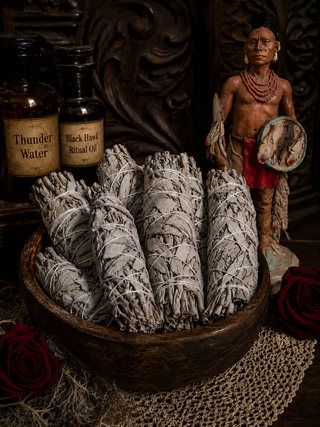 Bowl of sage smudge sticks with bottles labeled 'Thunder Water' and 'Black Ritual Oil', and a figurine in the background.