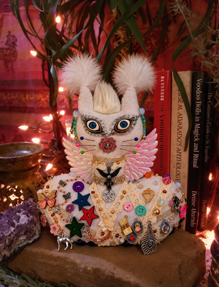 A highly detailed, handmade white cat figurine decorated with colorful beads, pins, feathers, and faux fur ears, styled on a stone surface next to books and warm string lights.