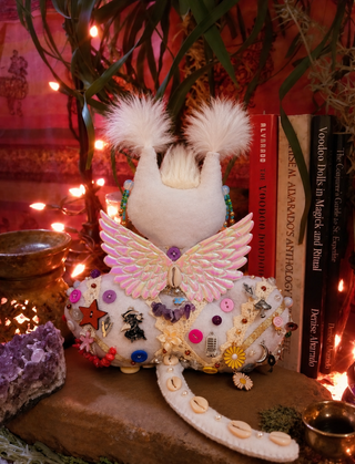 A whimsical handmade white felt creature doll with fluffy white feathers, pink embroidered wings, and a decorated white skirt adorned with buttons, beads, and shells, styled on a stone surface with books, crystals, and fairy lights.