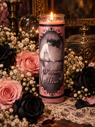 A lit pink glass candle with a vintage-style label reading 'Witching Hour,' surrounded by black and pink roses, white baby's breath, lace, and ornate dark metal decor.