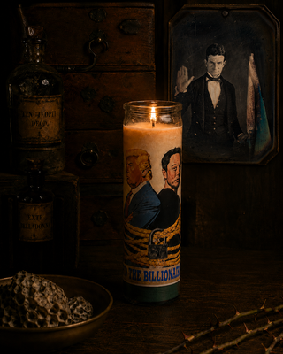 Candle with illustrated faces and text, surrounded by bottles and a painting in a dimly lit setting