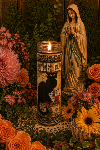 Candle with black cat design, statue of a religious figure, and flowers on a table.