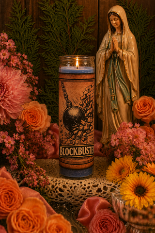Candle with 'Blockbuster' branding in front of a statue of a religious figure surrounded by flowers.