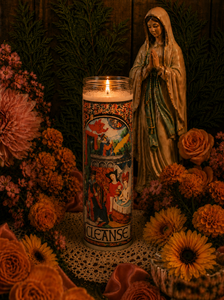 Candle labeled 'Cleanse' with a figurine and flowers on a dark background
